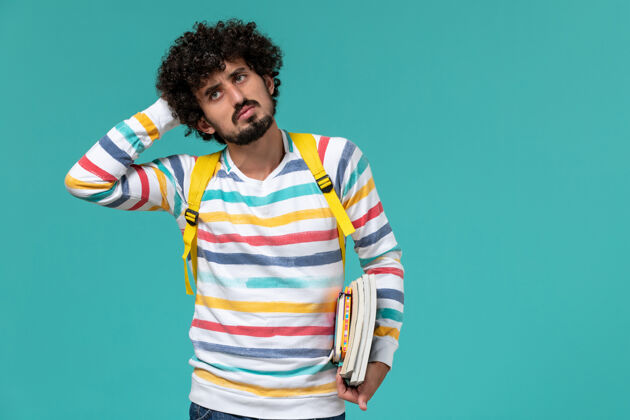 思考穿着条纹衬衫 背着黄色背包 拿着书 在蓝色墙上思考的男学生的正视图抱男学生学生