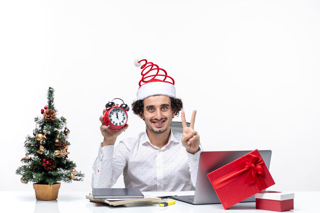 成人年轻快乐的商人戴着圣诞老人的帽子 拿着钟 在白色背景的办公室里展示两个时钟微笑年轻人