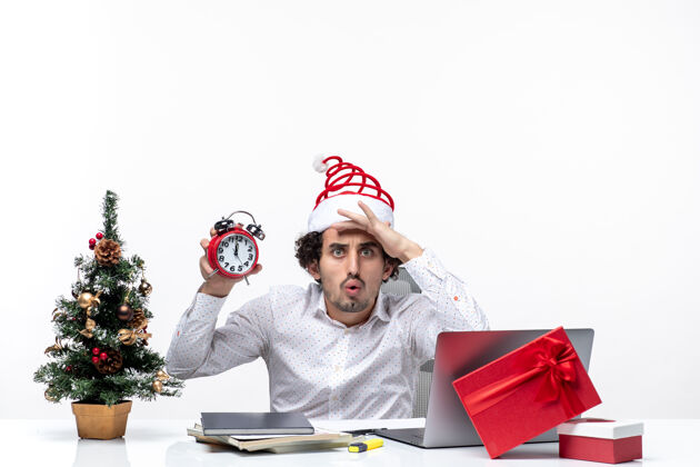 办公室戴着圣诞老人帽 拿着闹钟 头脑风暴的年轻人坐在白色背景的办公室里圣诞老人男性帽子