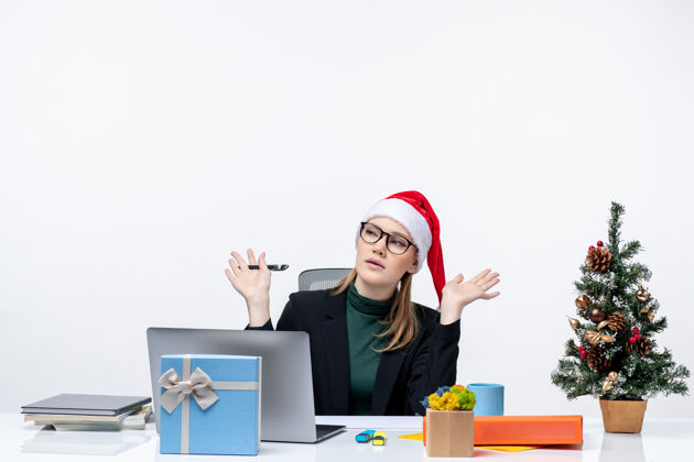 圣诞节困惑好奇的金发女人 戴着圣诞老人的帽子坐在一张桌子旁 桌子上放着一棵圣诞树和一份礼物 背景是白色的女商人圣诞树女人