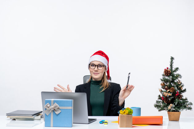 笔记本在白色背景的办公室里 一个戴着圣诞老人帽子的金发女人坐在桌子旁 桌子上放着圣诞树和礼物金发礼物女人
