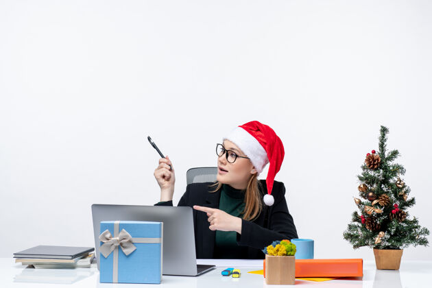 圣诞老人一个好奇的金发女人 戴着圣诞老人的帽子 坐在一张桌子旁 桌子上放着一棵圣诞树和一份礼物 看着左边白色背景上的东西坐着秘书电脑