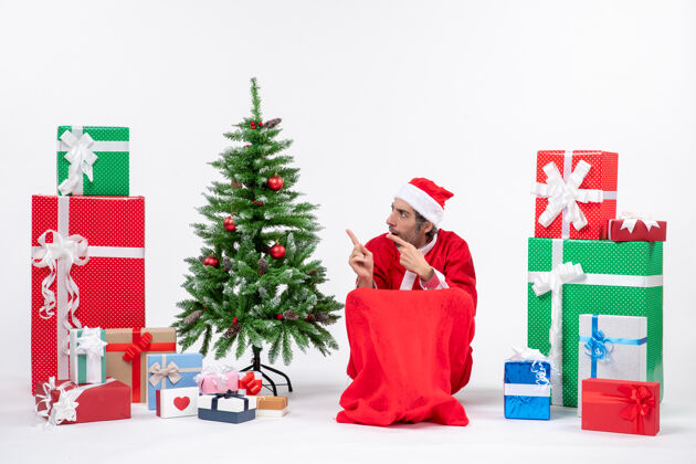 帽子一个悲伤的年轻人打扮成圣诞老人 拿着礼物和装饰过的圣诞树 在白色背景上指着右边的东西圣诞年轻人男人