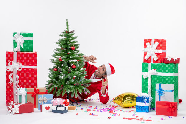 礼物圣诞气氛 年轻的圣诞老人指着自己躲在圣诞树后面的白色背景上不同颜色的礼物冬青指向圣诞老人