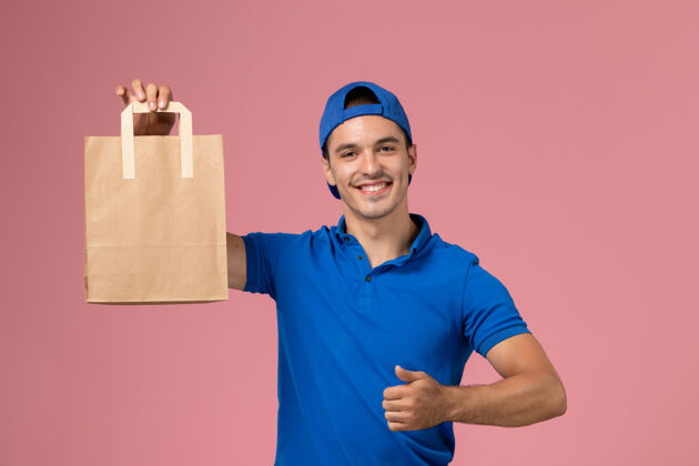 制服正面图：身穿蓝色制服和披风的年轻男性快递员 手上拿着快递纸包裹 微笑着站在粉红色的墙上服务男性年轻男性快递