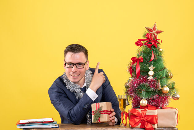 男人年轻人指着坐在圣诞树旁的桌子上的东西 在黄色的礼物上年轻人年轻人坐着