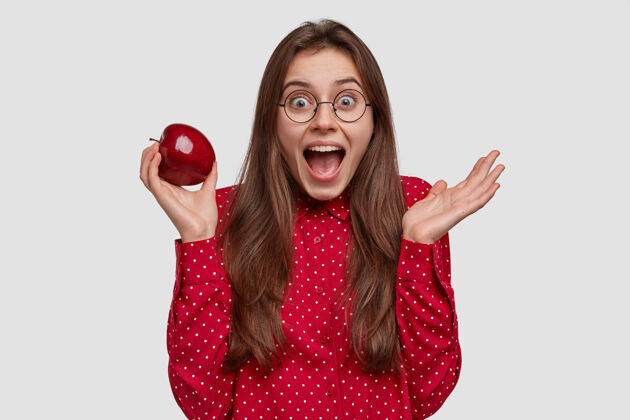 外观美女孤立镜头有愉快的表情 一手拿着红苹果 表达积极的情感 穿着正式的衬衫波尔卡多眼镜营养