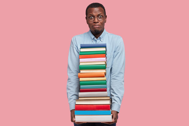 大学迷惑不解不满的黑皮肤男人拿着厚厚的一堆书 穿着正式的衬衫 模特们站在粉色的工作室墙上文学举行教育