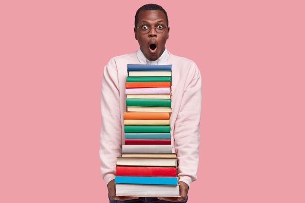 堆照片中穿着白色休闲套头衫的黑人年轻人提着一堆书 戴着圆眼镜 站在粉红色的墙上眼镜可怕教科书