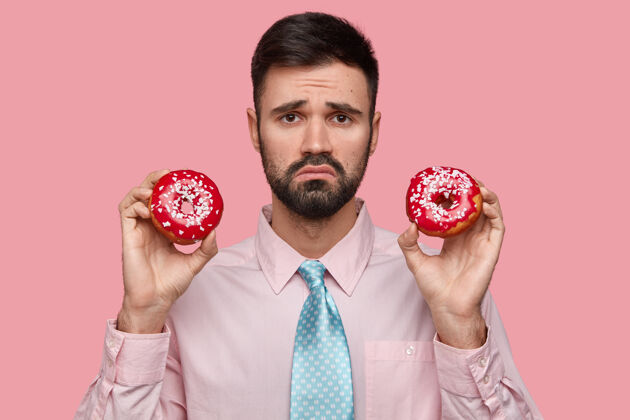 不高兴不高兴的男人留着深色的胡茬 皱着眉头 手里拿着两个美味的环形甜甜圈 不高兴得不能吃甜食不满领带外观