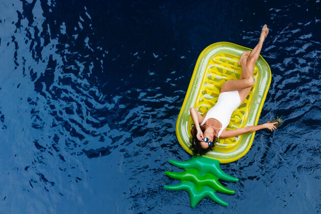 风景戴着太阳镜躺在菠萝床垫上的迷人女孩穿着泳衣的可爱棕褐色女模特在游泳池里放松的户外照片头顶泳装女性