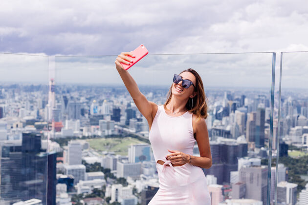 度假年轻快乐微笑的高加索女旅客 穿着合身的衣服 戴着墨镜站在曼谷的高楼上 手里拿着电话顶部玻璃地板