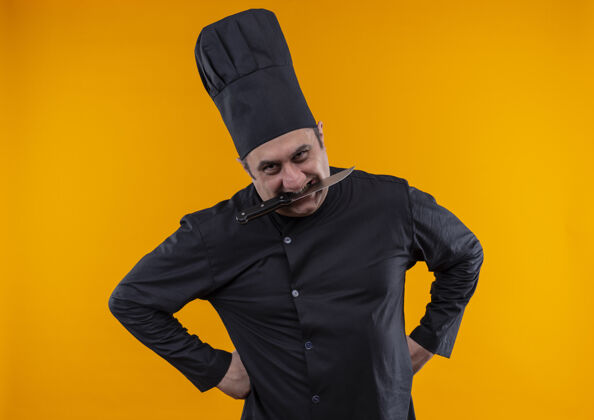中年高兴的中年男厨师穿着厨师制服 嘴里叼着刀站在黄色的墙上厨师刀厨师