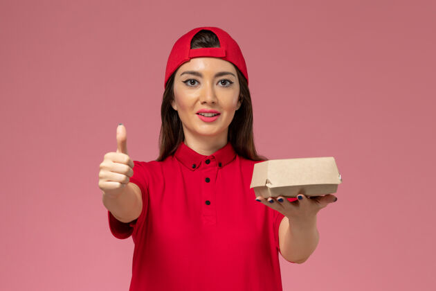 粉色正面图：身穿红色制服和斗篷的年轻女快递员 手上拿着一个小小的快递食品包 放在粉红色的墙上汉堡包裹人