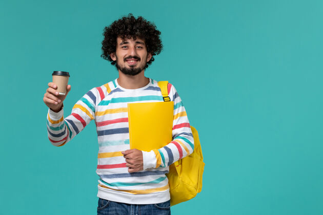 微笑穿着条纹衬衫 背着黄色背包 拿着文件和咖啡的男学生在蓝色墙上的正视图档案学校人