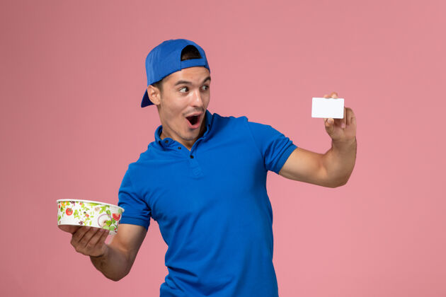 持有正面图身穿蓝色制服披肩的年轻男性信使手持白色卡片和浅粉色墙上的圆形投递碗碗圆快递
